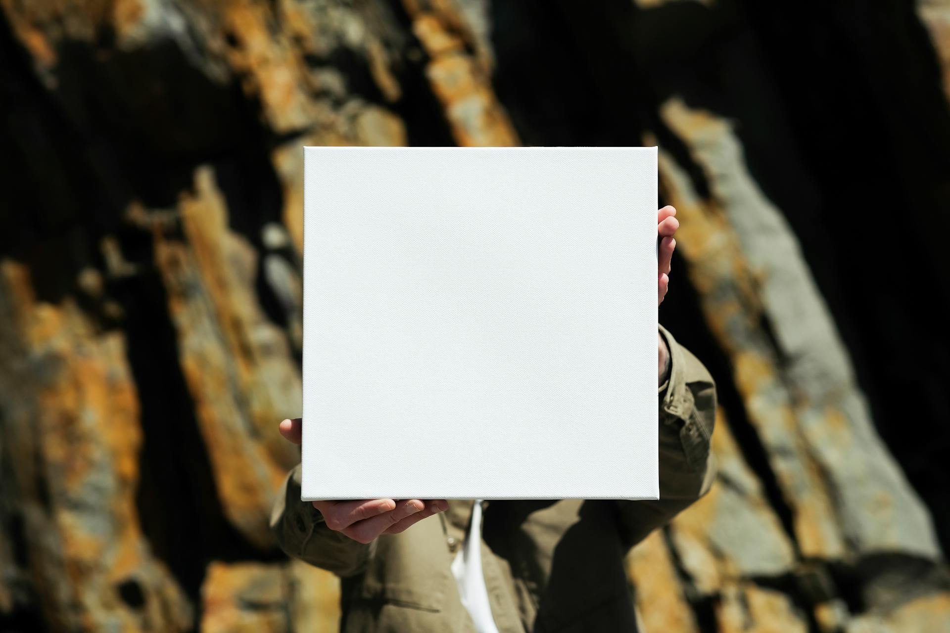 Hands presenting a blank white canvas against a rocky outdoor background, perfect for mockup designs.