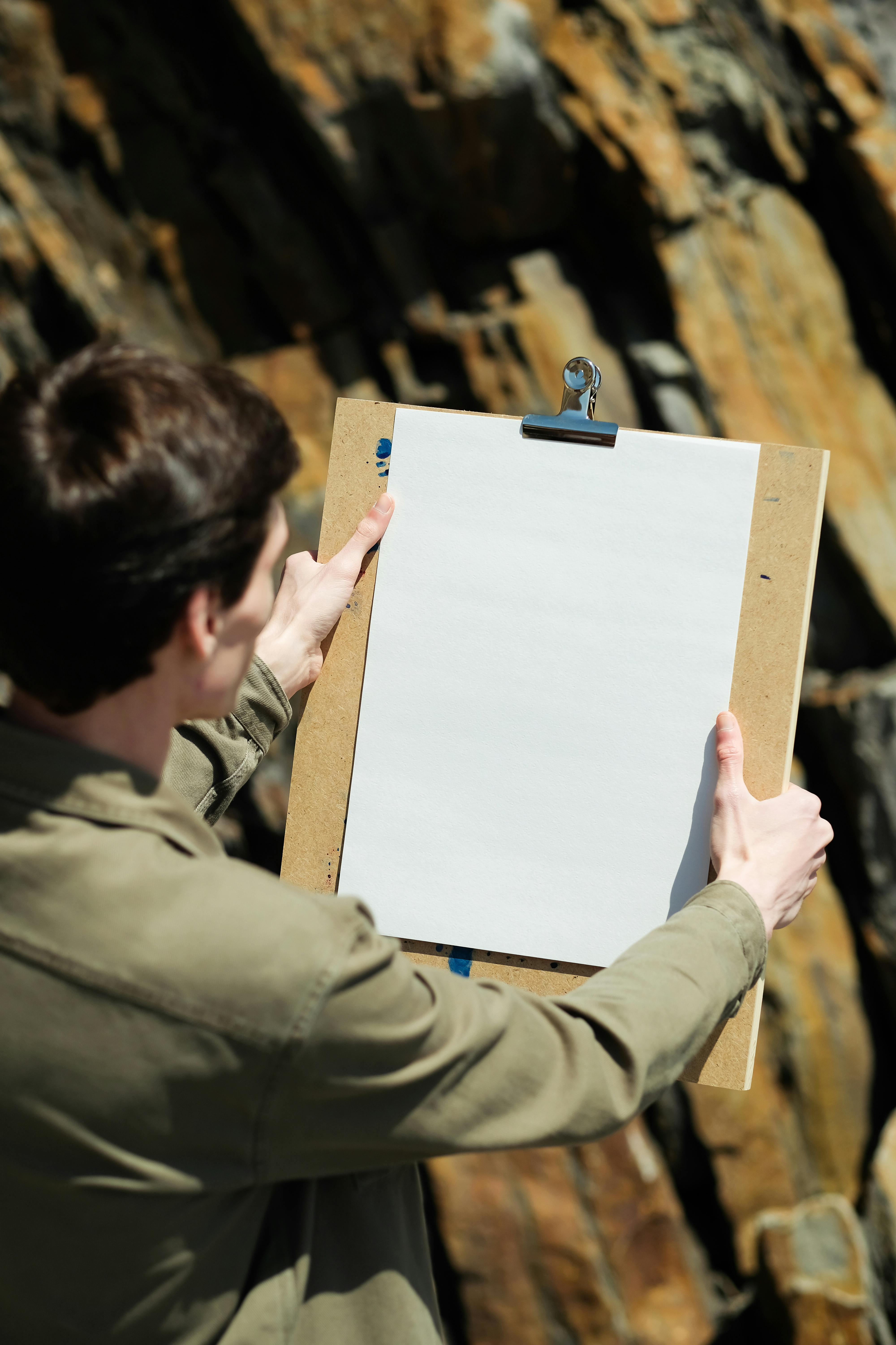 A back view of a man with short hair holding a blank clipboard against a rocky background.