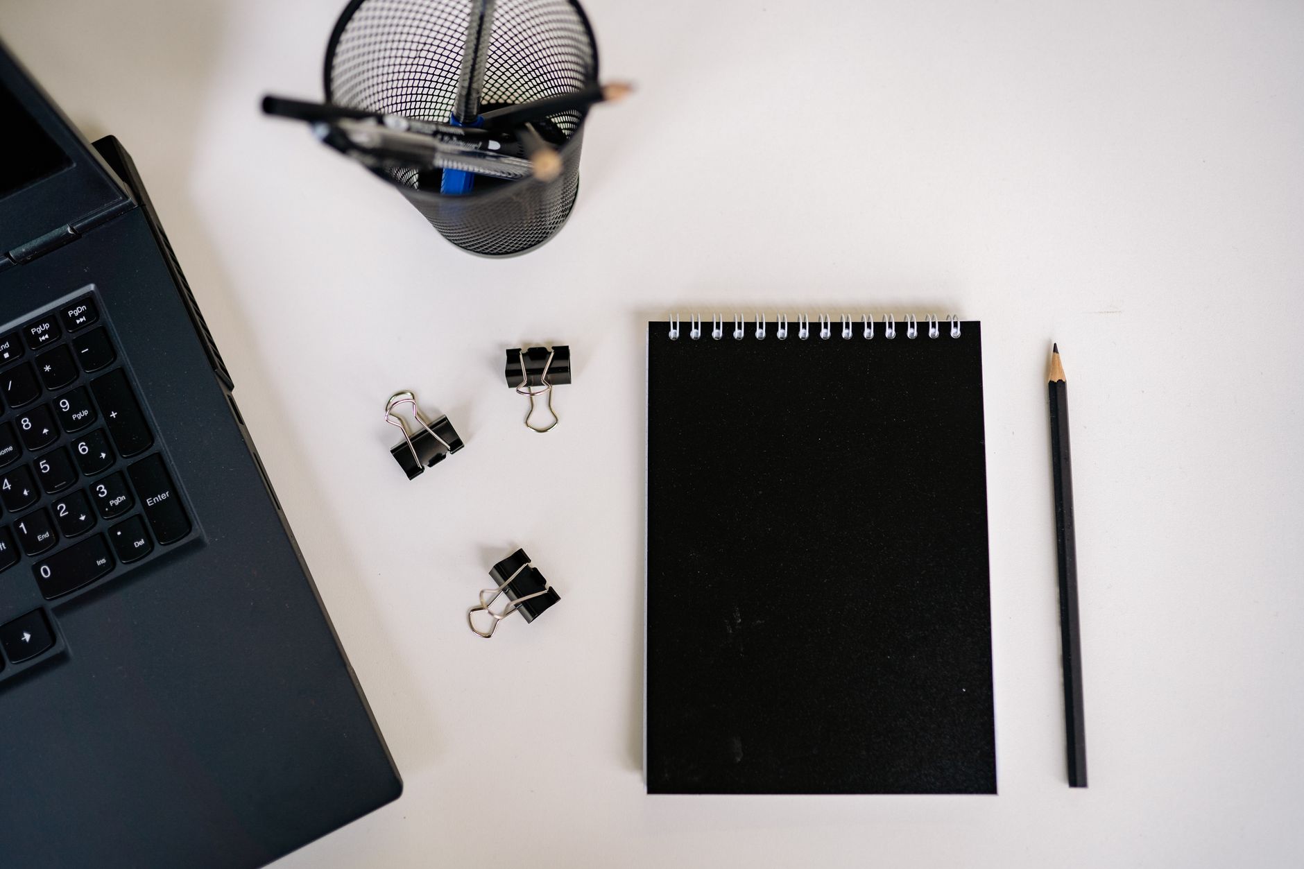 Overhead view of modern office supplies including black notepad, laptop, pencil, and clips.