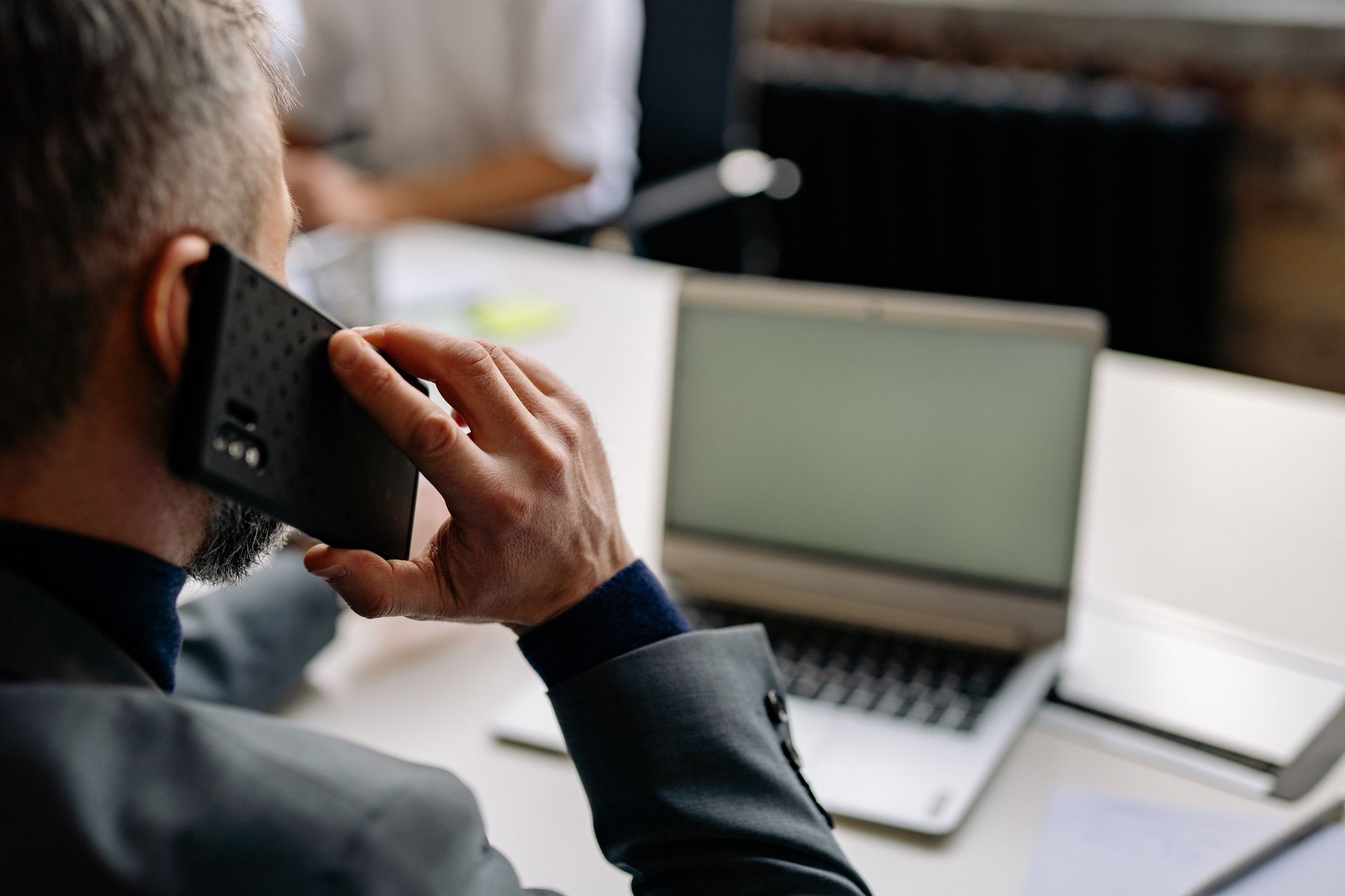 Professional man using cellphone in office setting, multitasking with a laptop.