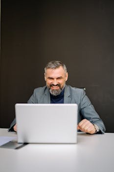 Smiling businessman in a gray suit using a laptop at a desk in an office setting.