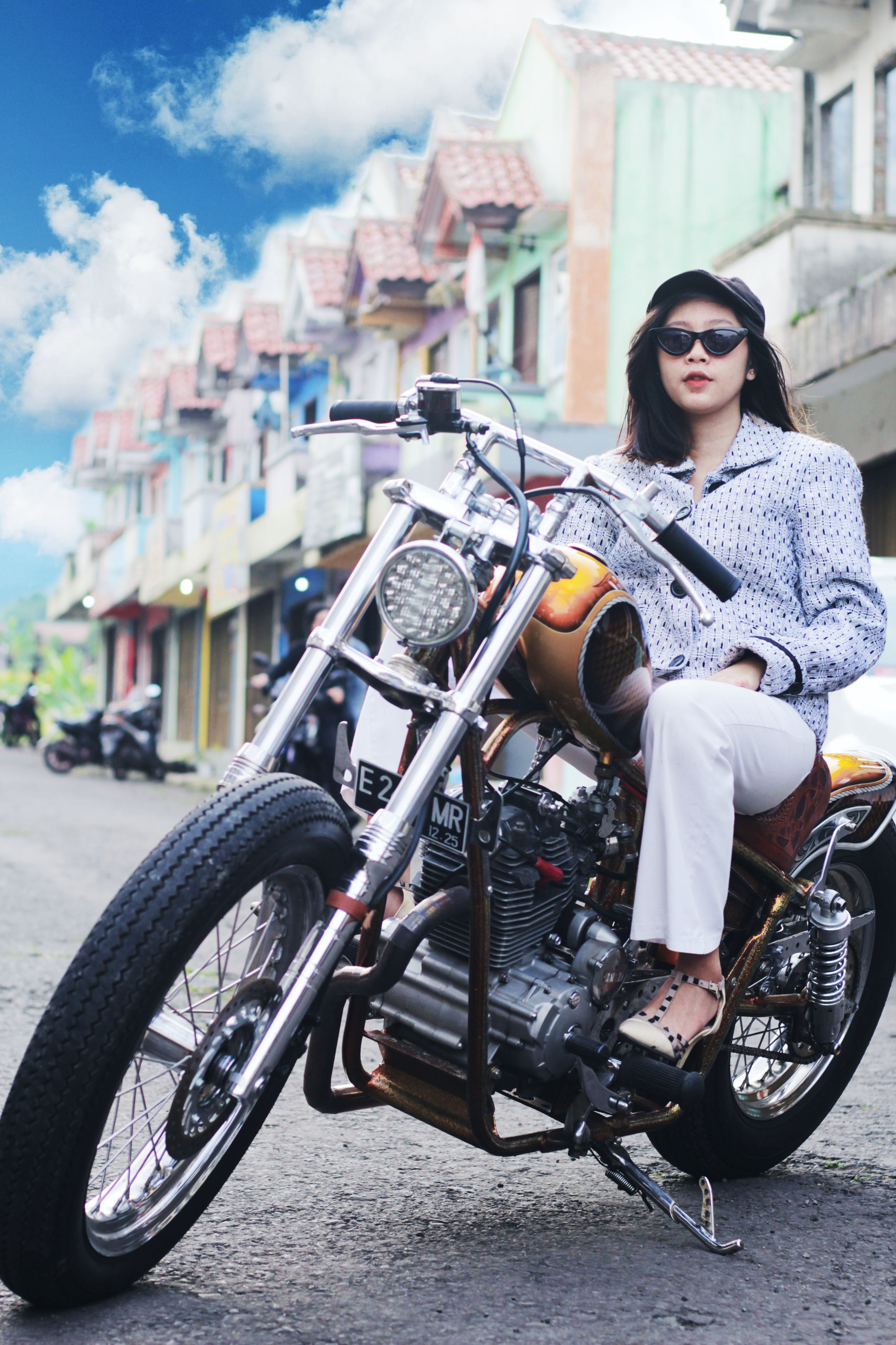 A young woman poses stylishly on a motorcycle in an urban street wearing sunglasses and fashion-forward attire.