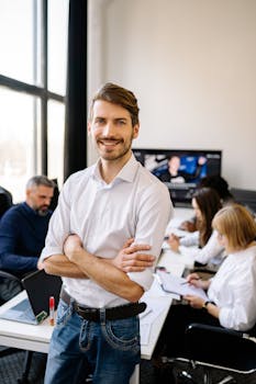Smiling man standing confidently in a modern office environment with colleagues working in the background.