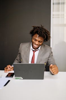 A cheerful African American man in a suit working on a laptop in a modern office setting.
