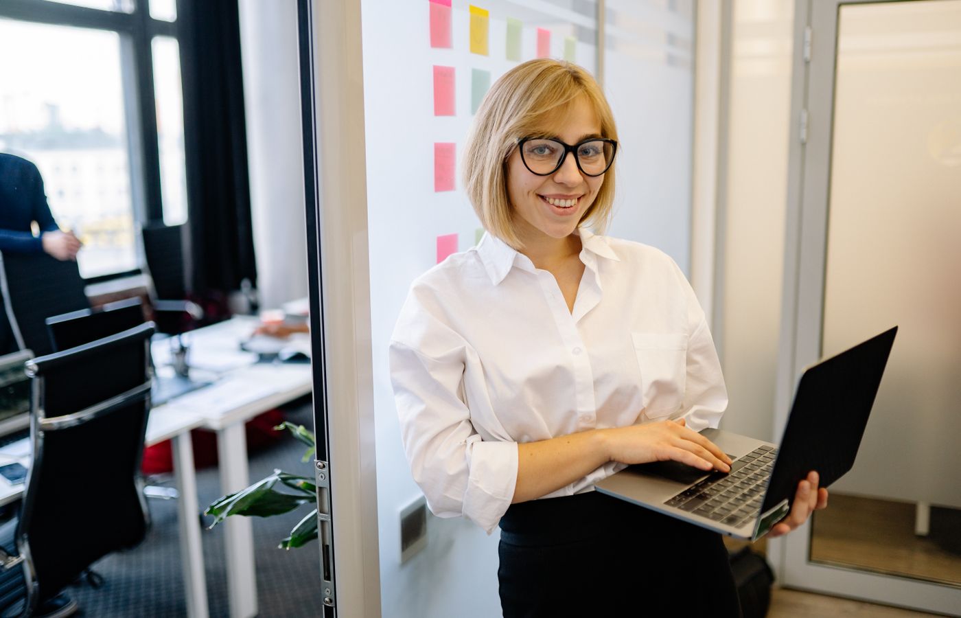 Young woman in glasses smiling confidently in a modern office setting with a laptop.