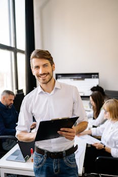 Professional man smiling while holding clipboard in modern office, colleagues working in background.