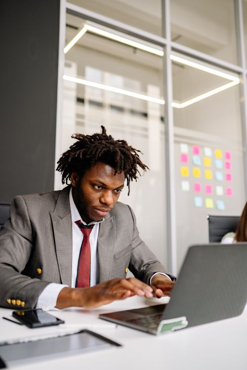 Free Black businessman in formal attire working intently on a laptop in a modern office setting. Stock Photo