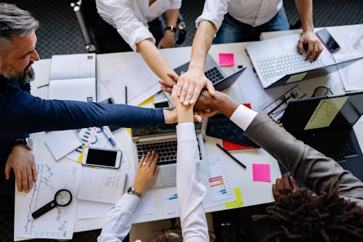 High-angle shot of diverse hands together over a desk with laptops and paperwork, symbolizing teamwork.