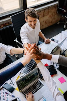 Group of diverse colleagues joining hands around a desk, symbolizing teamwork and unity.