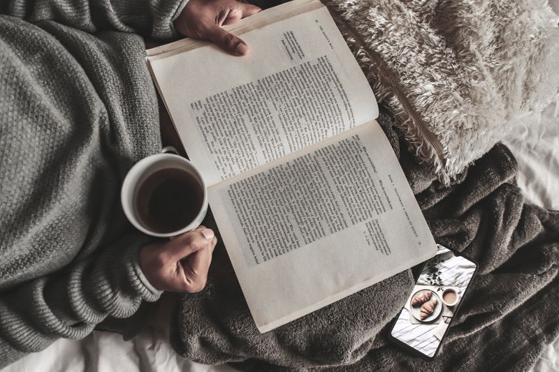 From above of crop anonymous female with mug of hot tea reading book under plaid near smartphone at home