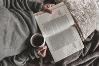 Top view of crop anonymous female sitting under warm plaid and reading book with cup of tea