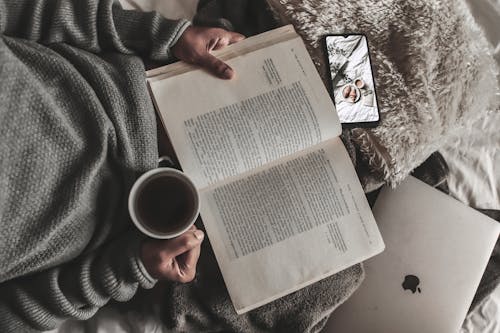 Free Top view of crop anonymous female reading novel placed on soft plaid near laptop and cellphone while drinking tea Stock Photo