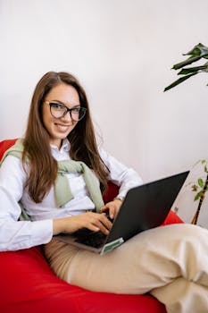 Confident young woman smiling with a laptop on a cozy chair, working remotely.