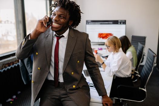 Confident businessman with dreadlocks smiling while on a phone call in a modern office setting.