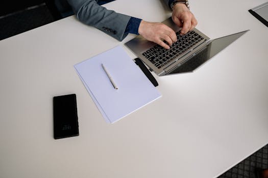 Top view of businessman typing on a laptop at a neat white desk with phone and papers.