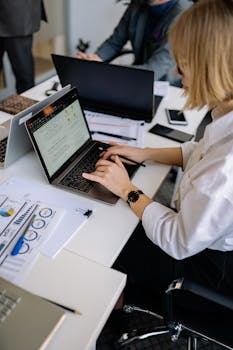 Woman working on laptop at office desk with documents and graphs in view.