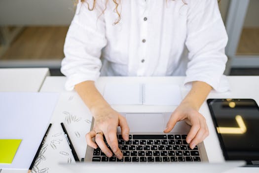 Close-up of woman's hands typing on laptop keyboard in bright, modern office space. Ideal for work and technology themes.