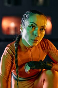 A cosplayer in striking orange sportswear kneeling, with intense gaze, colorful lighting.