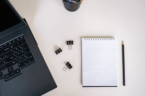 Free Top view of an office workspace featuring a laptop, notepad, pencil, and binder clips on a clean desk. Stock Photo