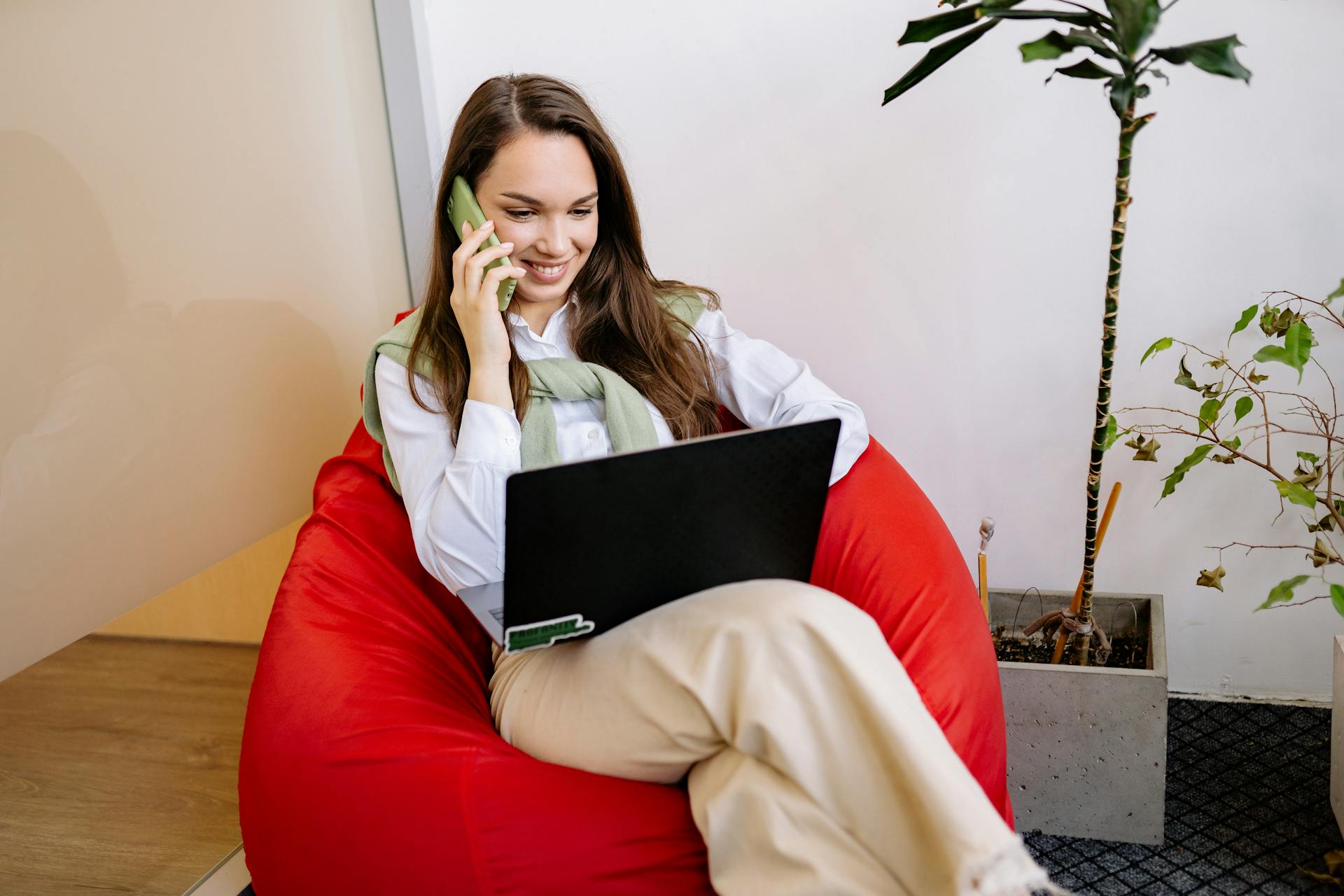 A woman sits on a bean bag working remotely with a laptop and talking on the phone.