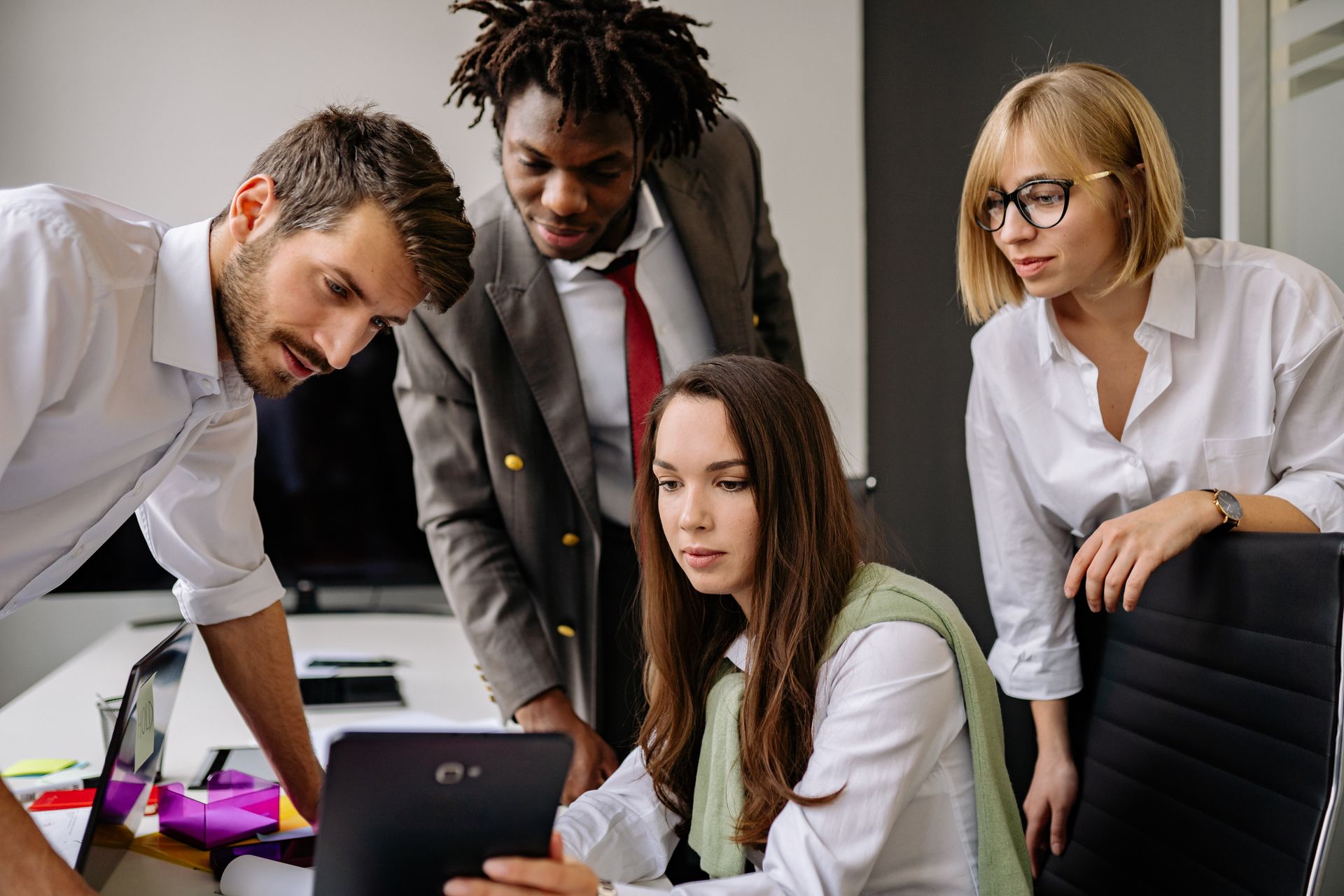 A diverse group of colleagues engaged in team discussion, utilizing a tablet in a modern office setting.
