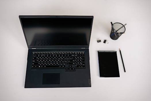 Flat lay of a laptop, notepad, pencil, and office supplies on a white desk.