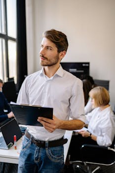 A bearded man in a white shirt and denim pants holding a clipboard in a busy office setting.