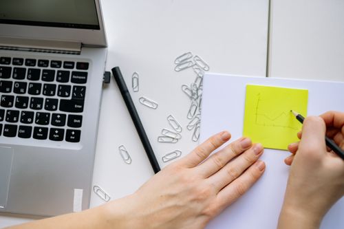 Free A person sketches a graph on a sticky note next to a laptop and paper clips, suggesting planning and creativity. Stock Photo