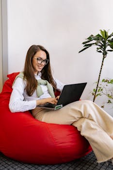 Young woman with eyeglasses smiling while typing on a laptop from a red bean bag chair indoors.