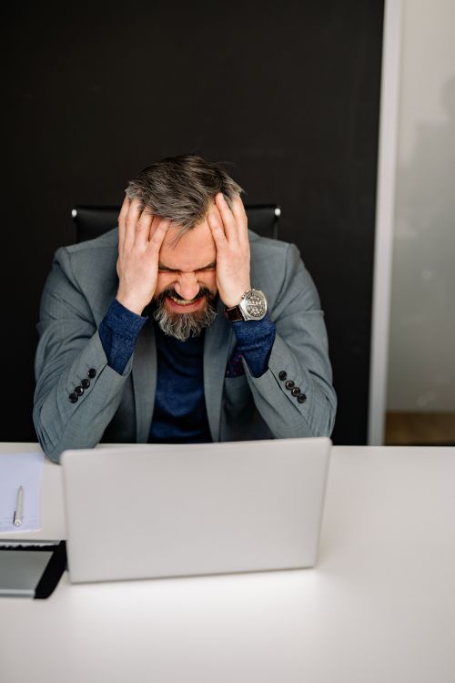 Free Stressed businessman in gray suit with head in hands at office desk, facing laptop. Stock Photo