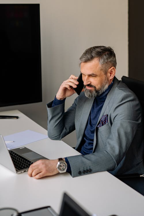 Free Man in Gray Suit Sitting on a Chair in front of His Laptop while Having a Phone Call Stock Photo
