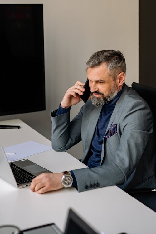 Man in Gray Suit Sitting on a Chair in front of His Laptop while Having a Phone Call