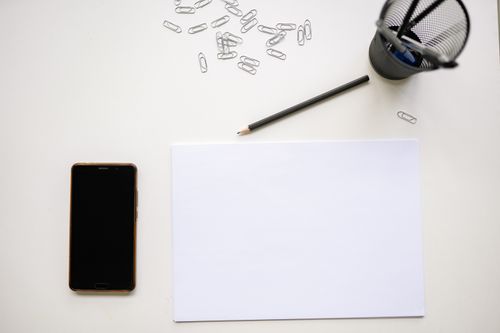 Free Top view of a minimalist desk layout with smartphone, pencil, paper clips, and stationery essentials on white paper. Stock Photo