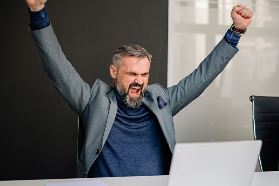 Thrilled businessman with raised arms celebrating success at his office desk.