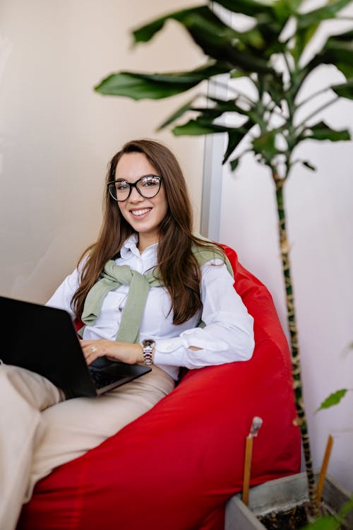 Woman in White Dress Shirt Sitting on a Bean Bag Chair Using Black Laptop Computer