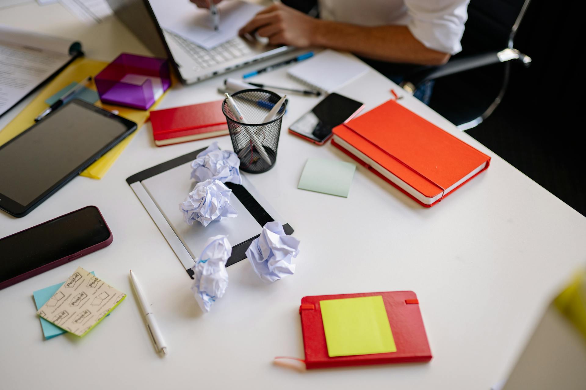 A cluttered office desk with crumpled papers, notebooks, and stationery.