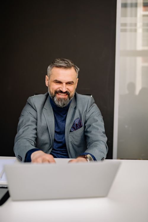Free Confident businessman with beard working on laptop, smiling in modern office. Stock Photo