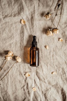 Brown glass bottle with pipette on fabric surrounded by dried flowers, top view.