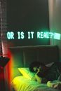 Young lady lying in bed in dark room with neon illumination and Or Is It Real inscription on wall