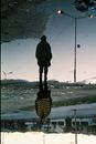 Upside down of young pensive female in warm clothes and hat standing in puddle at roadside and looking away in town at sunset