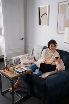 Adult woman in sweater using smartphone and laptop on sofa, creating a cozy tech-friendly vibe.