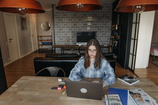 Young woman in eyeglasses working from home on a laptop, wearing a plaid shirt.
