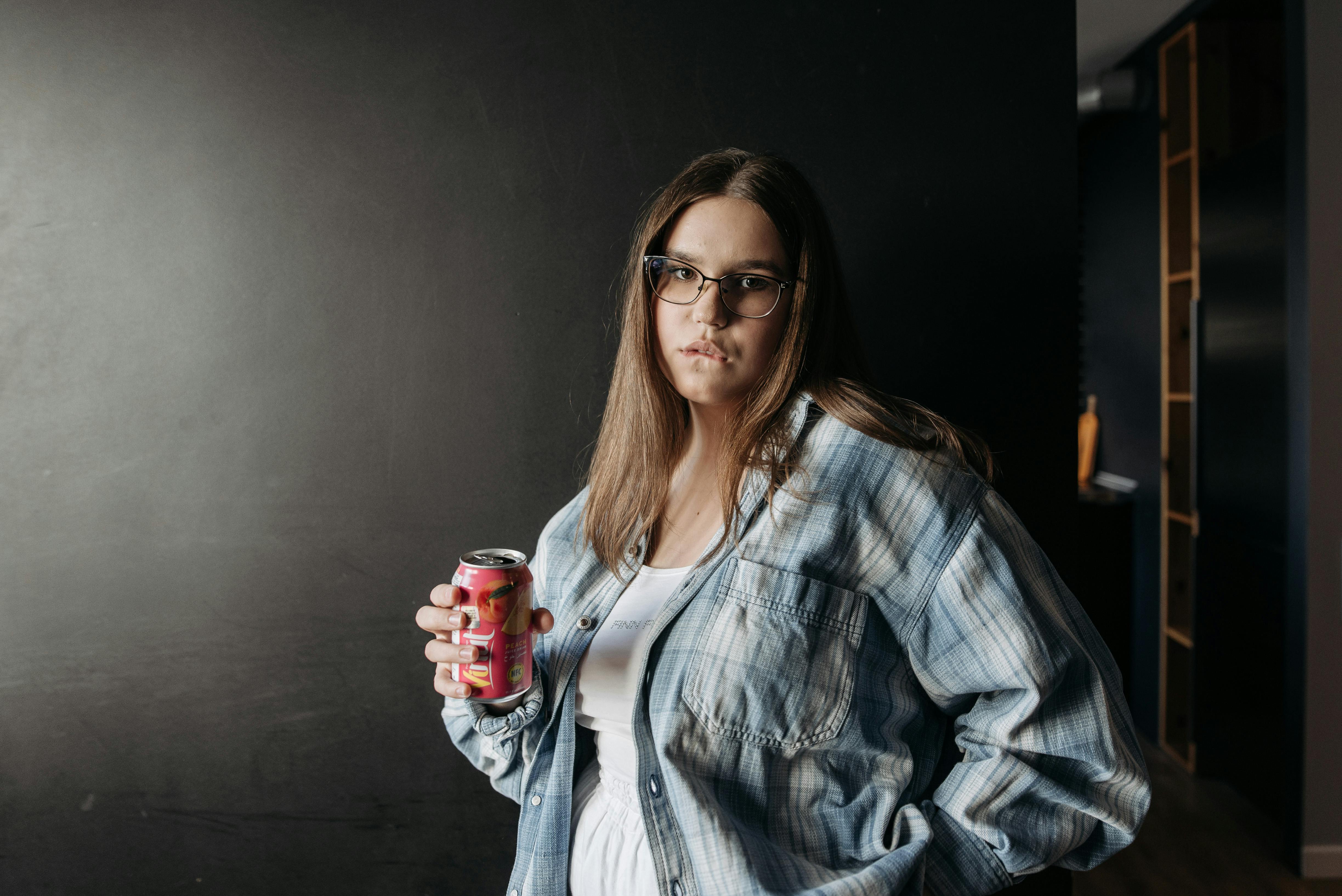 Woman casually holding a soda can indoors, wearing plaid shirt and eyeglasses.