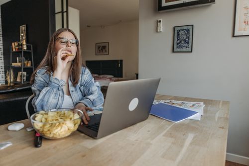 Free Woman eating chips while studying online at home, multitasking with laptop. Stock Photo