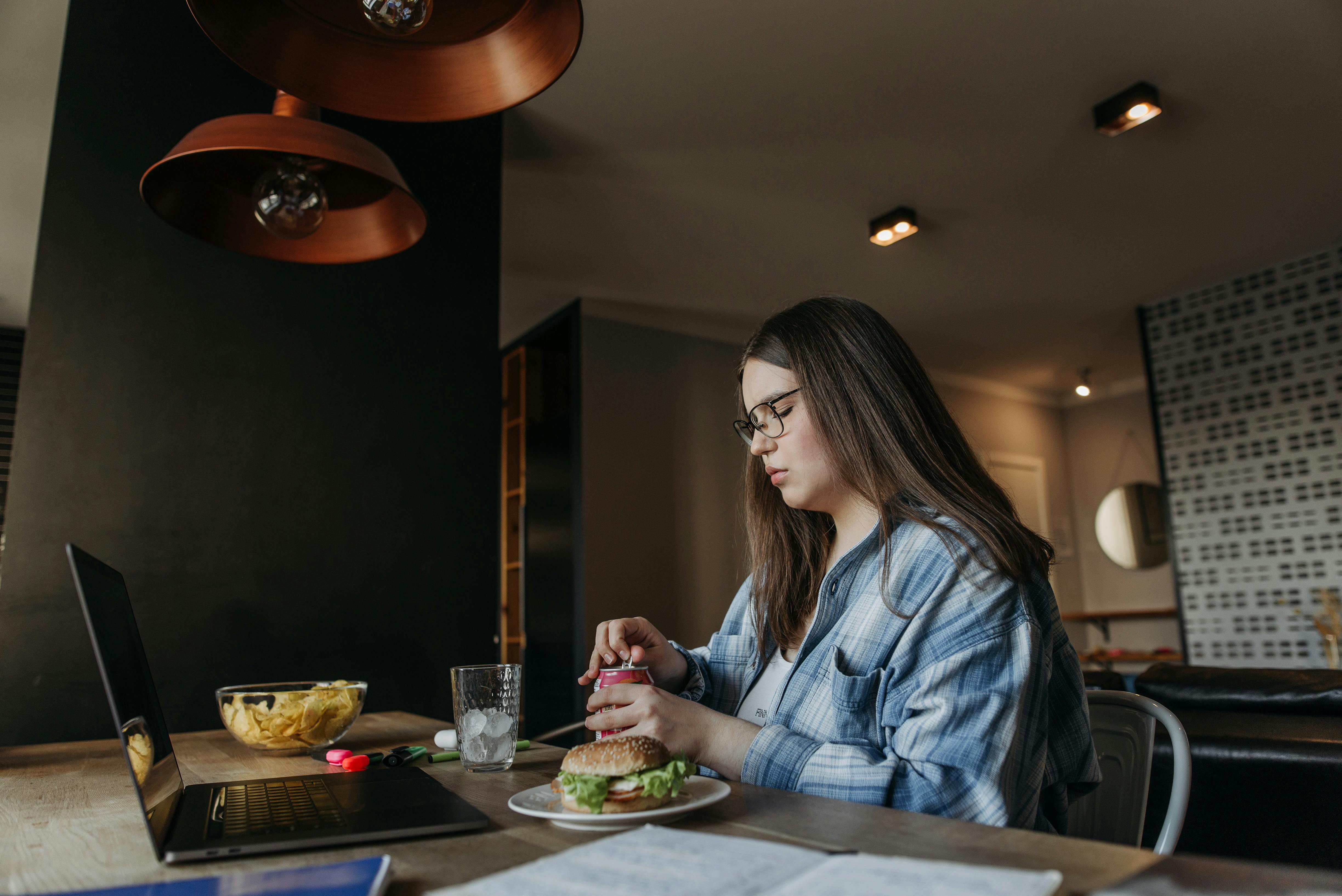 Woman eating lunch at home while working on laptop, featuring chips and a burger on a wooden table.
