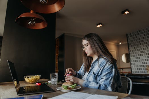 Woman eating lunch at home while working on laptop, featuring chips and a burger on a wooden table.