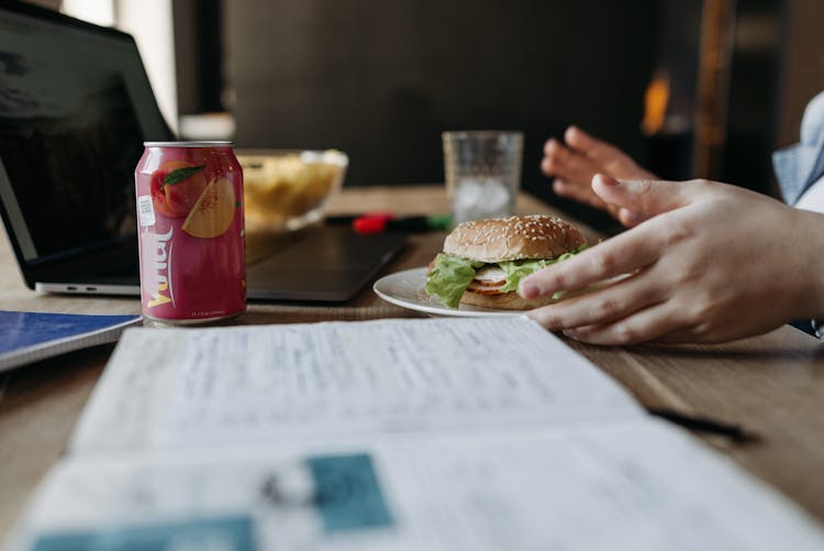 Person Sitting At A Desk With A Hamburger, Can Of Drink, And A Laptop