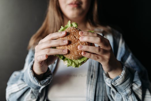 A person in a casual shirt holding a fresh burger with lettuce and sesame bun.