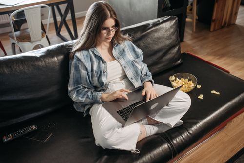 Free Woman working on laptop in casual setting at home with snacks on sofa, depicting remote work lifestyle. Stock Photo