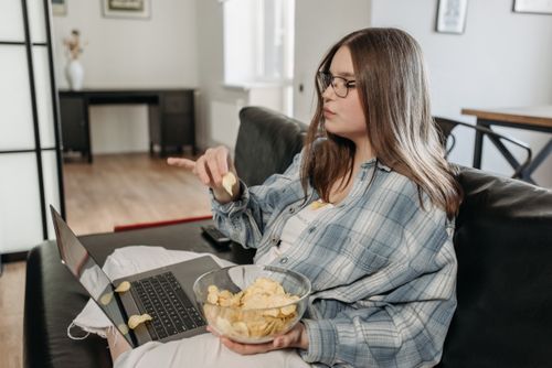 Free Woman in casual attire relaxing on a couch with a laptop and bowl of chips. Stock Photo